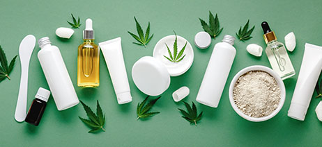 Assortment of THC lotions and serums in cosmetic bottles, surrounded by cannabis leaves, all on top of a green background