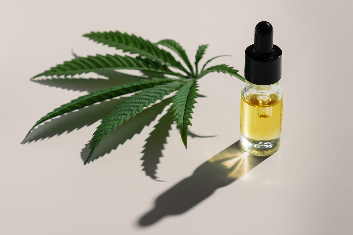 Clear cannabis tincture bottle with yellow liquid inside, sitting next to a cannabis fan leaf